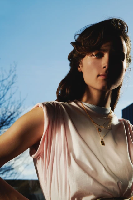 Portrait of a stylish young woman with layered gold necklaces wearing a pastel pink t-shirt against a bright blue sky backdrop.