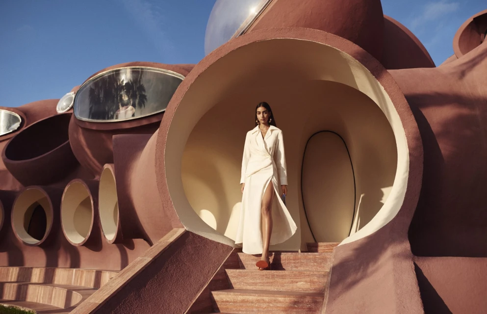 Adult woman in a white coat stands in front of an architectural, dome structure under a blue sky during the day.