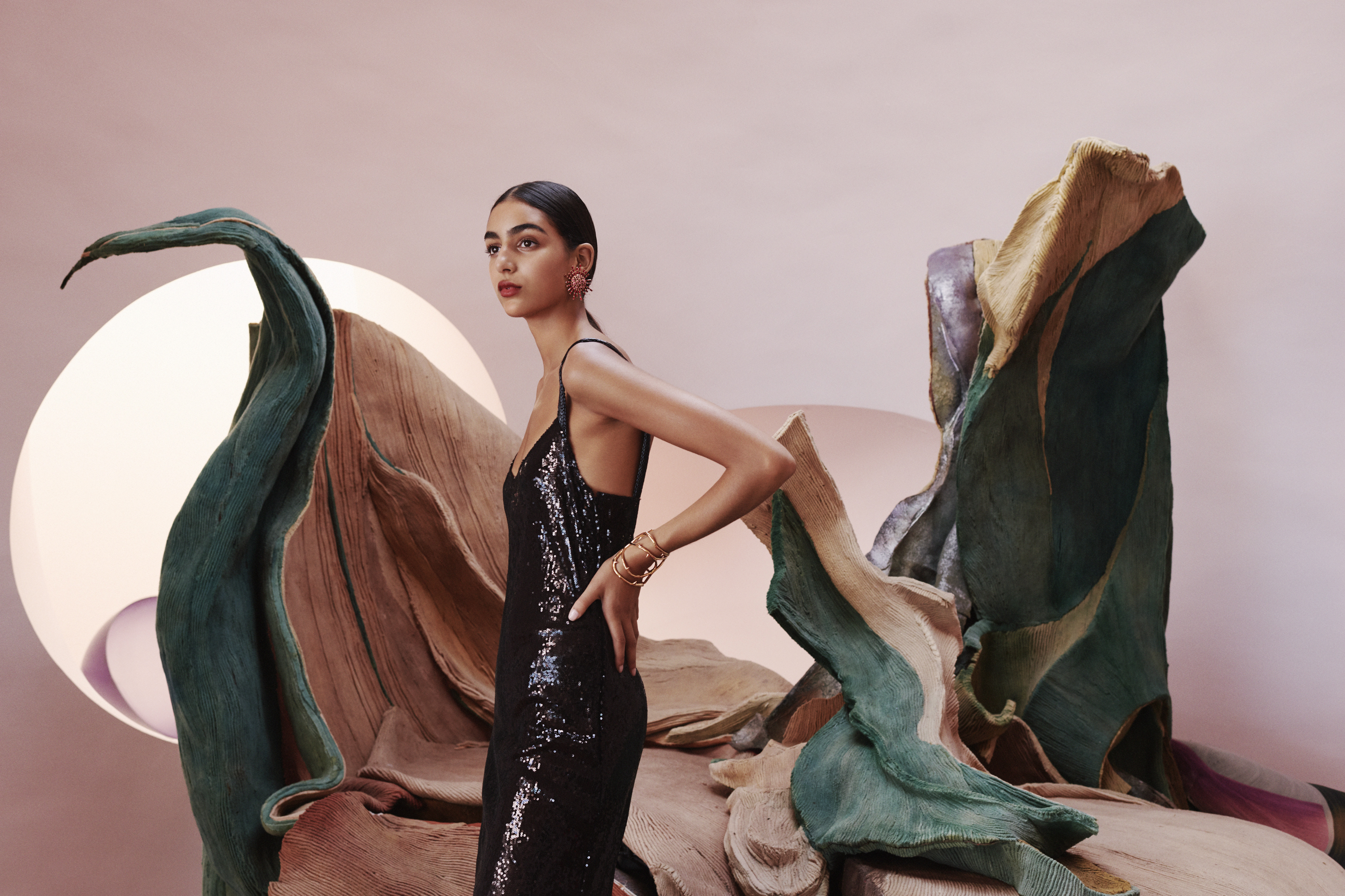 Elegant woman in a black sequin dress posing in front of textured, abstract sculptural art pieces against a muted pink backdrop.