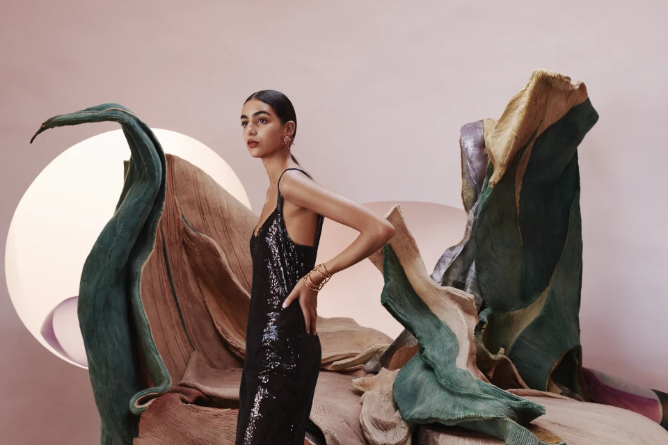 Elegant woman in a black sequin dress posing in front of textured, abstract sculptural art pieces against a muted pink backdrop.