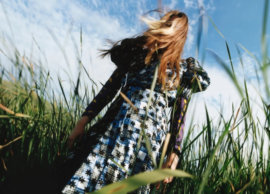 A woman in a patterned dress stands in a field of tall grass under a partly cloudy sky, viewed from a low angle emphasizing the sky.
