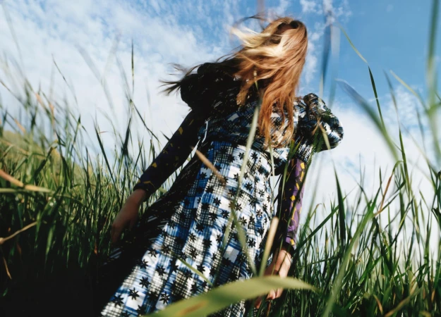 A woman in a patterned dress stands in a field of tall grass under a partly cloudy sky, viewed from a low angle emphasizing the sky.