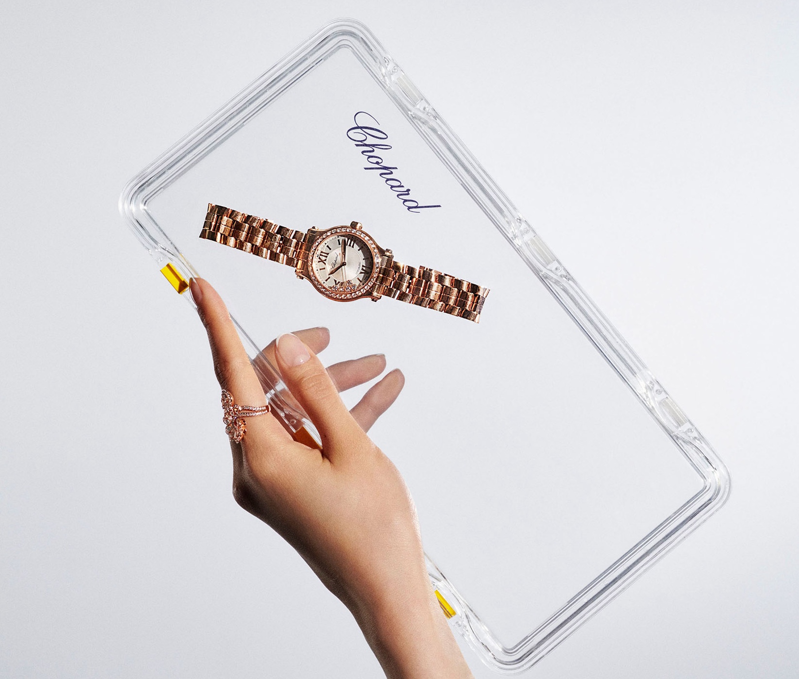 Rose gold wristwatch on a transparent display held by a woman's hand with a rose gold ring, set against a plain white backdrop.