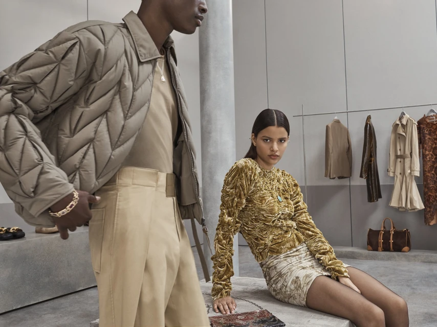 Two fashion models in modern apparel pose in an indoor studio. A man stands left in a quilted jacket while a woman sits on a stone bench.