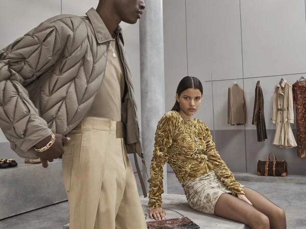 Two fashion models in modern apparel pose in an indoor studio. A man stands left in a quilted jacket while a woman sits on a stone bench.