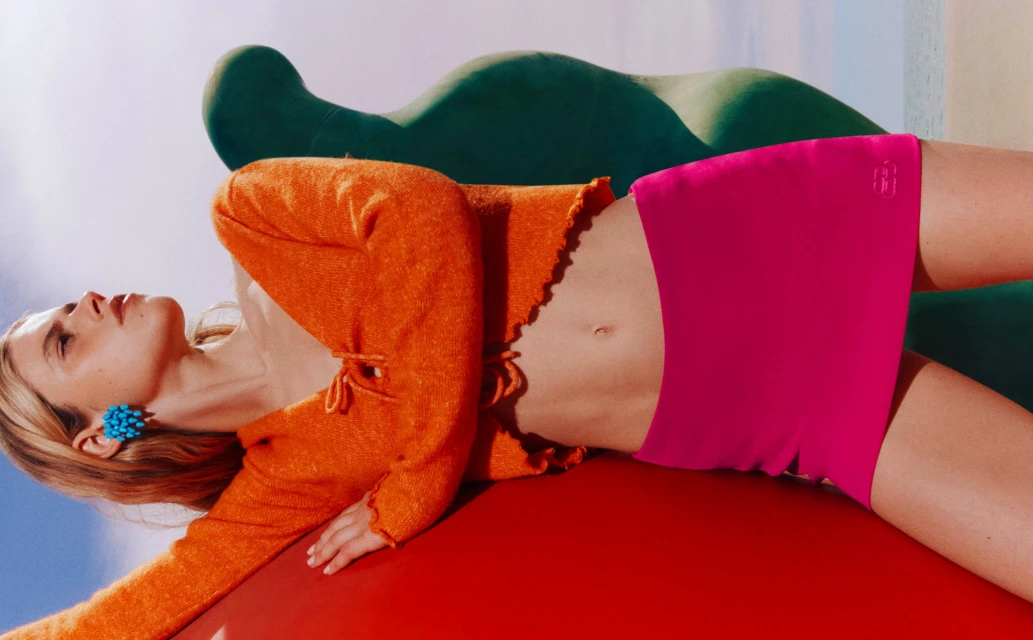 A fair-skinned young adult woman poses reclining with an orange sweater, pink miniskirt, blue earrings, against a colorful backdrop.