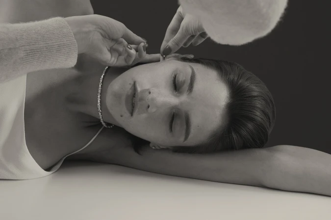Woman lying down with eyes closed while hands fit a delicate earring, wearing a glittering tennis necklace and white camisole.