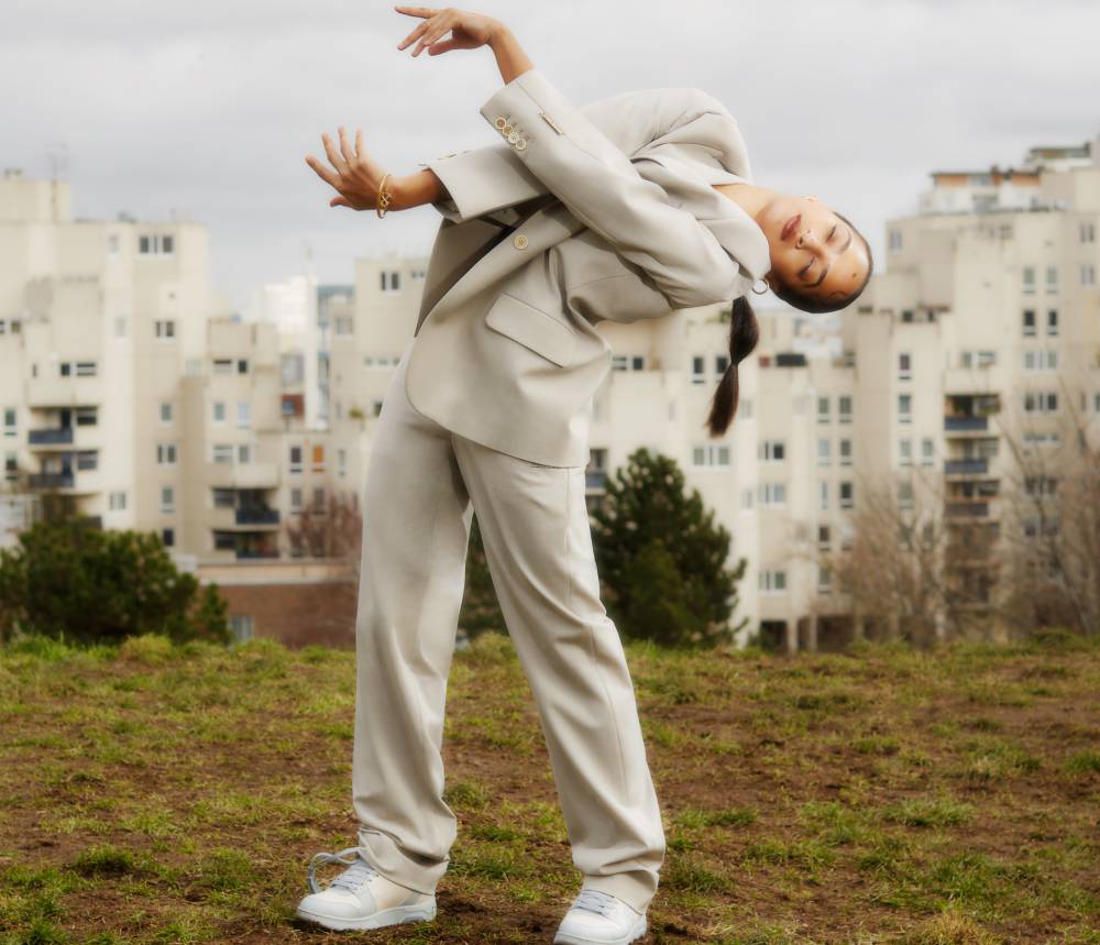 A woman in a beige suit poses on a grassy hill with buildings in the background, showcasing fashion and urban photography.