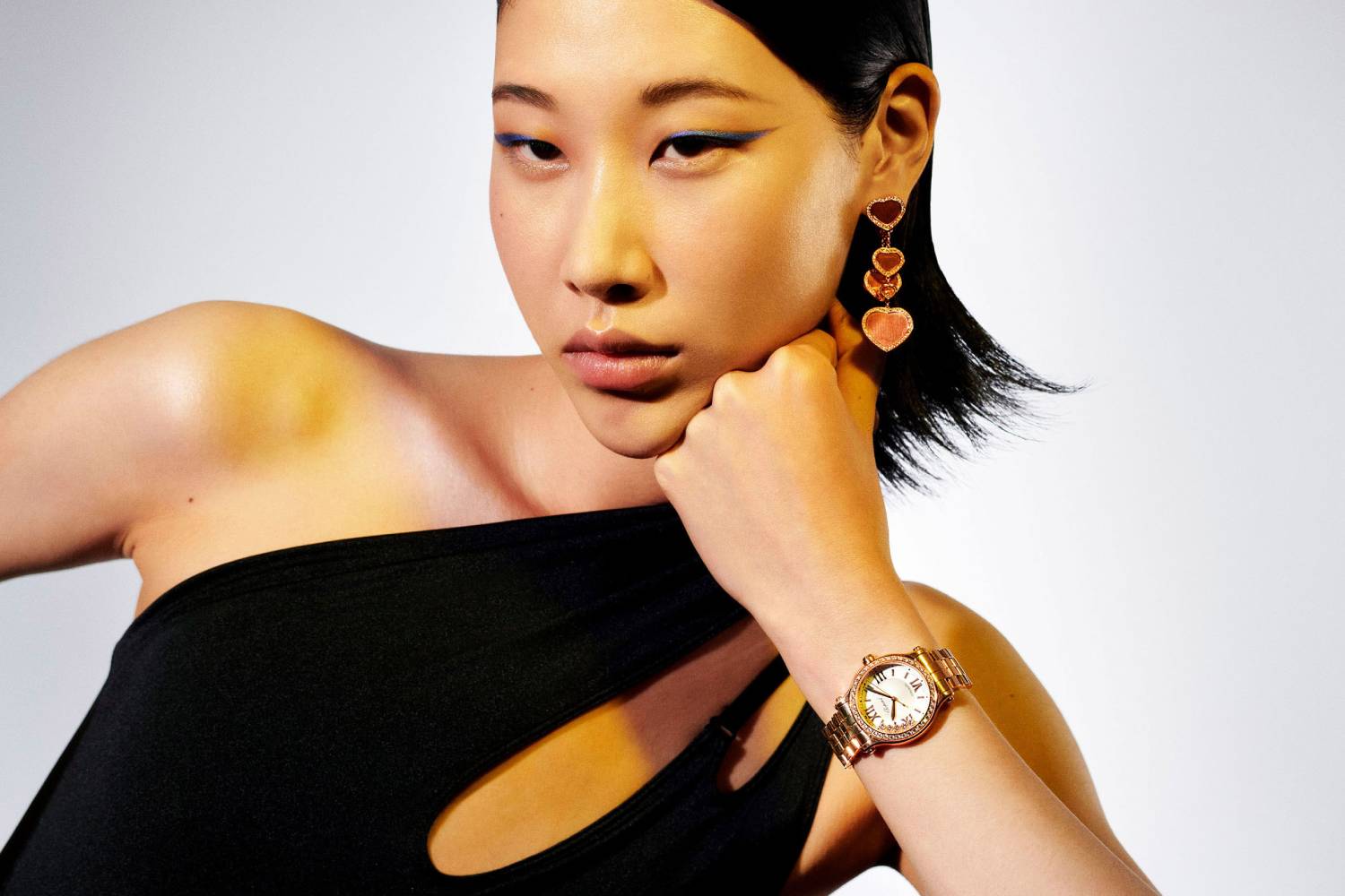 Close-up shot of an Asian woman wearing heart earrings and a wristwatch, posing in a black top against a clean, neutral background.