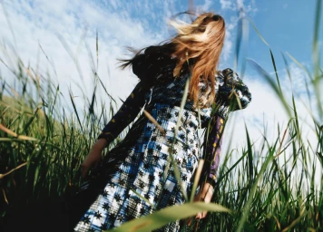 Low-angle view of a woman in a blue and white patterned dress standing in tall green grass with windblown hair against a blue sky.
