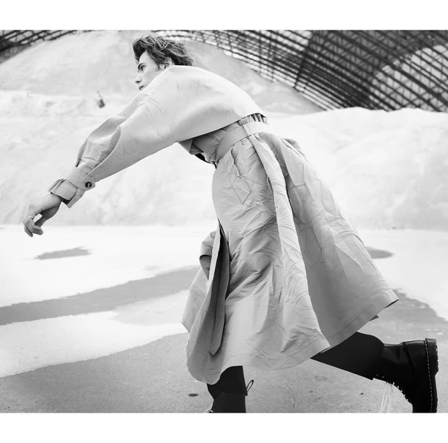 Monochrome fashion shot of a slender male adult striding forward wearing a draped jacket, skirt, and boots in a stark indoor location.