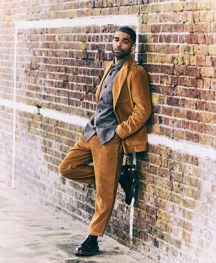 An adult male wearing a corduroy suit leans casually against a brick wall in an urban setting, showcasing fashion and style.
