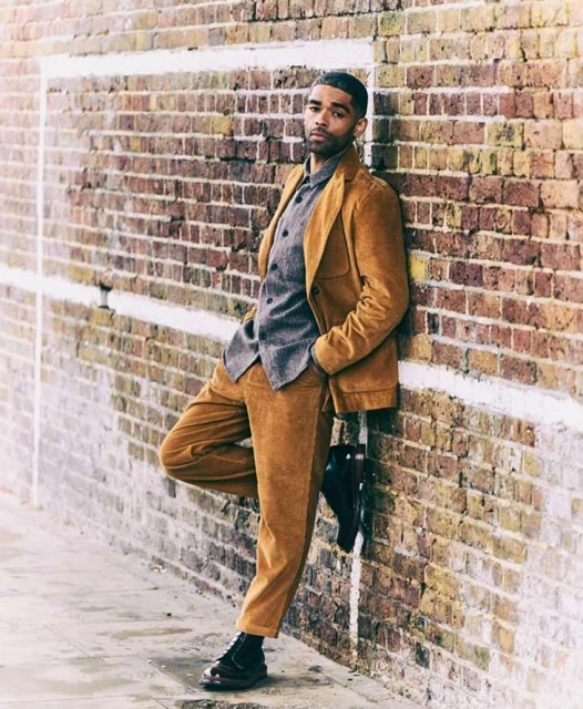 An adult male wearing a corduroy suit leans casually against a brick wall in an urban setting, showcasing fashion and style.