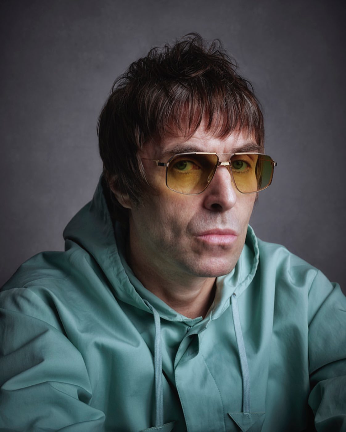 Close-up portrait of a man with brown hair, wearing yellow tinted glasses and a light blue hooded jacket against a gray background.