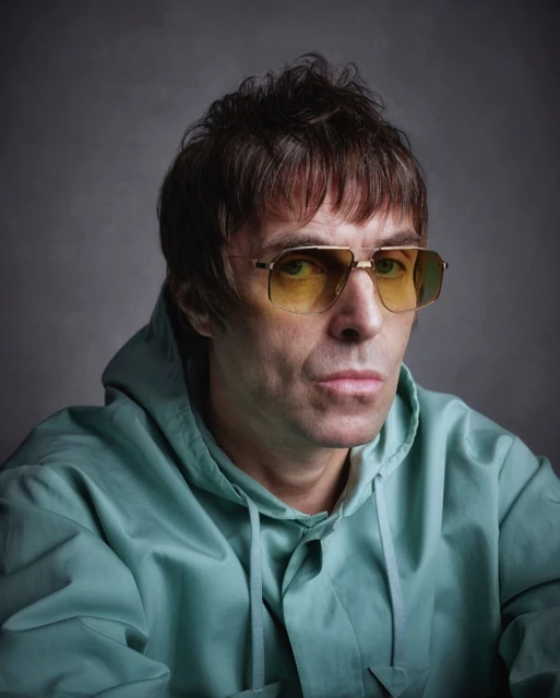 Close-up portrait of a man with brown hair, wearing yellow tinted glasses and a light blue hooded jacket against a gray background.
