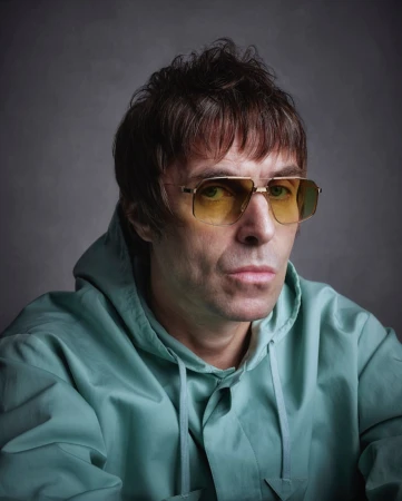 Close-up portrait of a man with brown hair, wearing yellow tinted glasses and a light blue hooded jacket against a gray background.