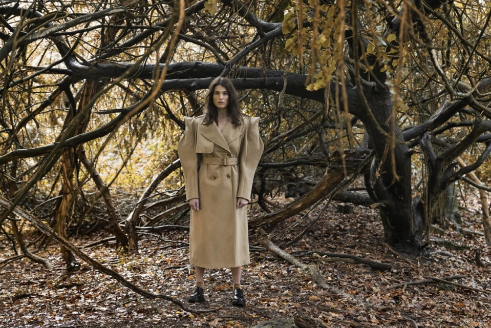 Woman in trench coat standing in a forest with bare branches and golden leaves, fashion photography, outdoor setting, autumn aesthetic.