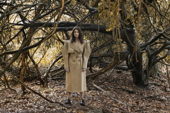 Woman in trench coat standing in a forest with bare branches and golden leaves, fashion photography, outdoor setting, autumn aesthetic.