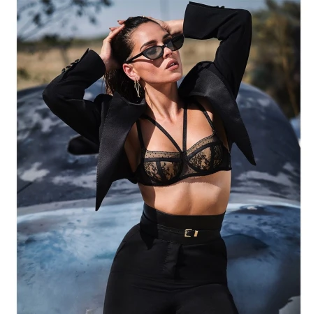 Woman wearing black bra, black trousers, and sunglasses posing outdoors with hands behind her head against a blurred natural background.