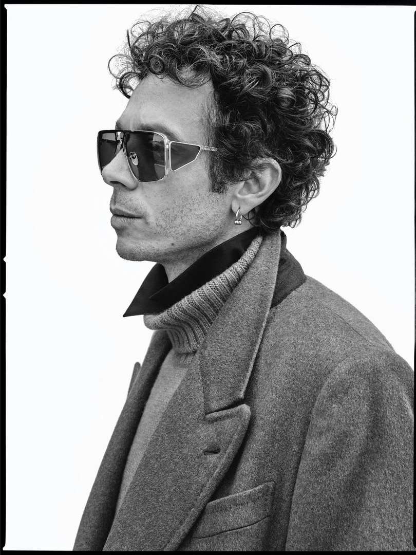 Profile portrait of a man with curly hair wearing oversized sunglasses, a turtleneck, and a grey coat against a plain white background.