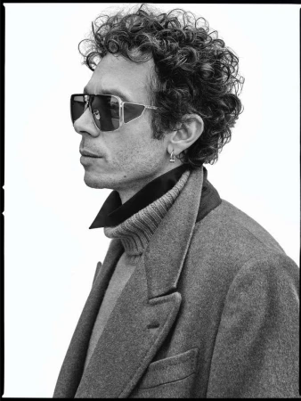 Profile portrait of a man with curly hair wearing oversized sunglasses, a turtleneck, and a grey coat against a plain white background.