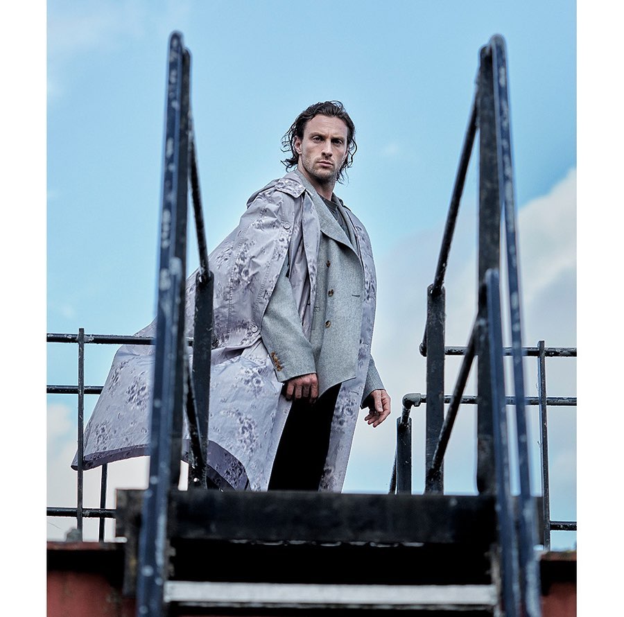 Man in patterned coat and suit standing on a stairway against a cloudy sky, looking at the camera in an outdoor setting.