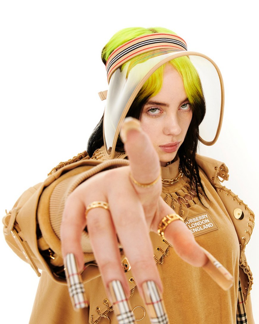 Close-up studio shot of a woman with blonde and black hair wearing a tan visor, gold jewelry, and a Burberry top with long nails.