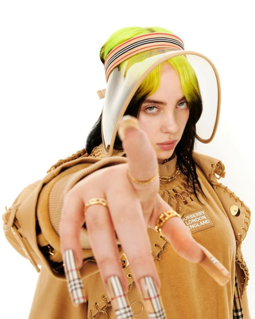 Close-up studio shot of a woman with blonde and black hair wearing a tan visor, gold jewelry, and a Burberry top with long nails.