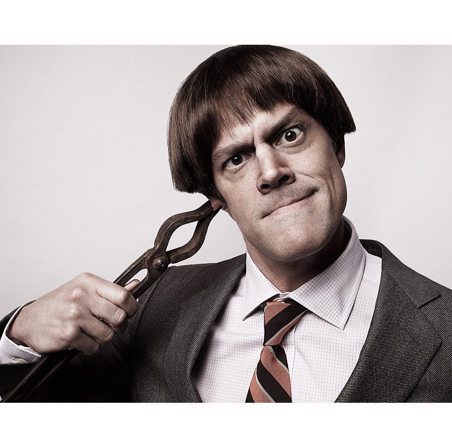Adult man with a dark bowl cut hairstyle wearing a gray suit, white shirt, and striped tie holding metal tongs to his ear against a gray background.