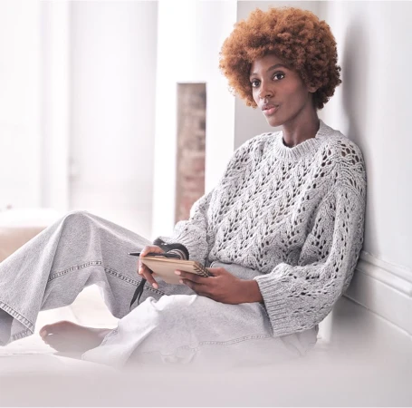 Black woman with short curly brown hair wearing a grey knit sweater and light wash jeans holding a small notebook