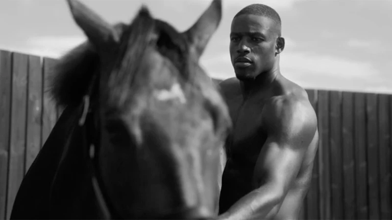 Black and white portrait of a shirtless black man standing beside a dark horse in front of a wooden fence by Gavin Bond.
