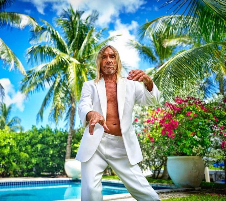Iggy Pop, wearing a white suit, dances outside in a tropical setting near a pool with palm trees and colorful flowers.