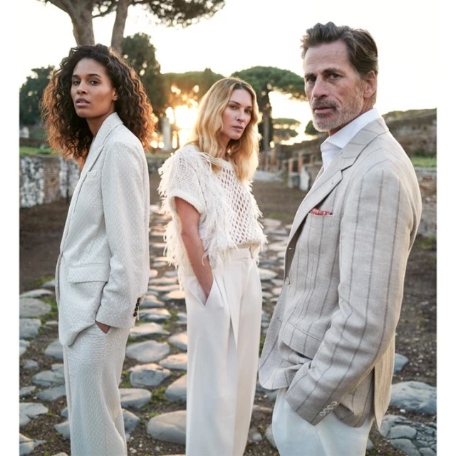 Three fashion models pose outdoors near Roman ruins in neutral-toned linen and knitwear. Sunlight creates a warm, editorial aesthetic.