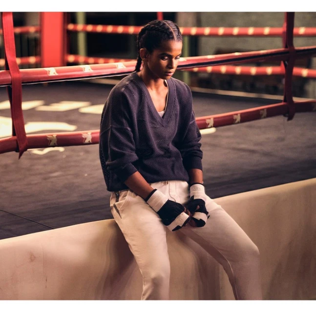Female boxer in sports attire pauses in the ring, looking down, with boxing ring in soft focus, neutral color grading, indoor lighting.