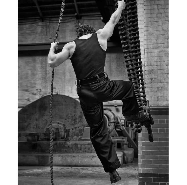 Monochrome shot of muscular man in tank top and trousers climbing a large metal chain in an industrial setting, back to camera.