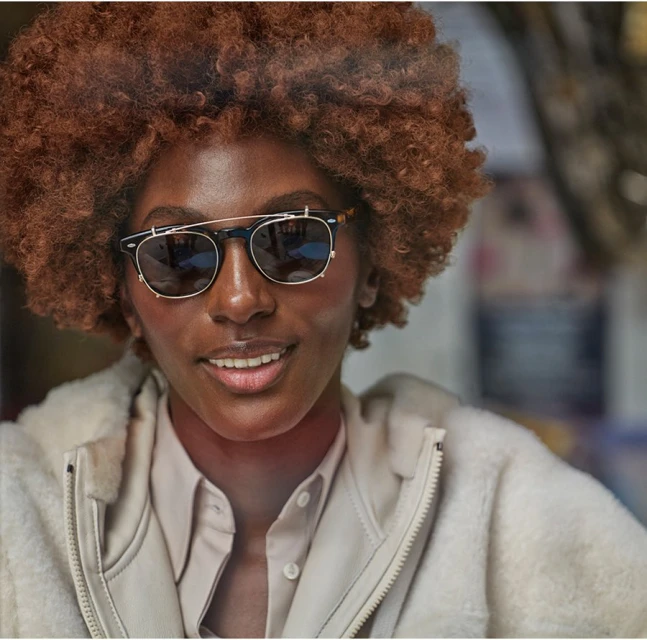 Close-up portrait of an African-descent adult female with auburn curly hair, stylish sunglasses, neutral make-up, and a light-color jacket.