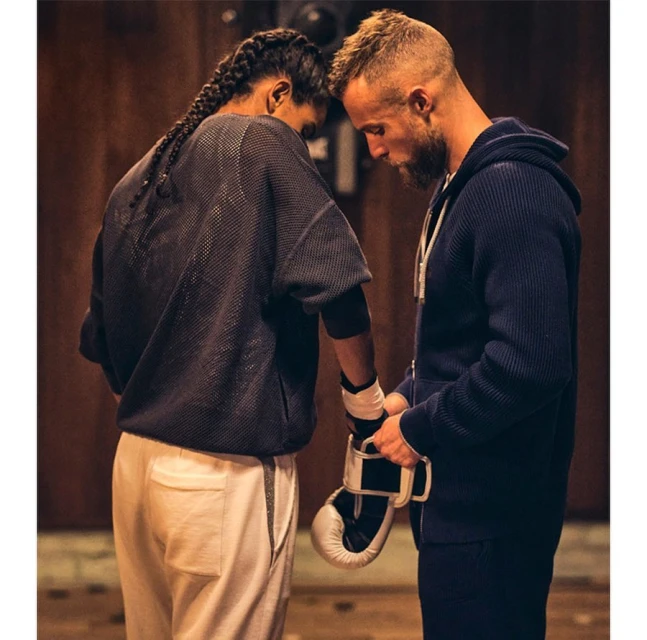 Two athletes face each other indoors. One wraps the other's hands with boxing tape, both dressed in athletic wear, with wood background.