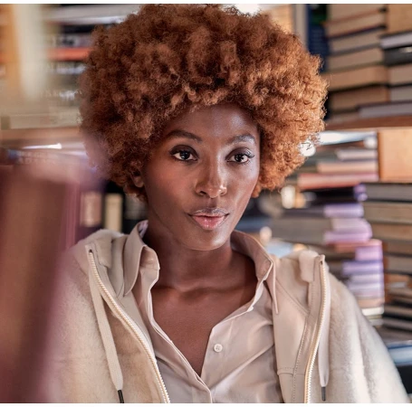 Portrait of a Black woman with voluminous curly ginger hair wearing a collared shirt and light jacket in a library