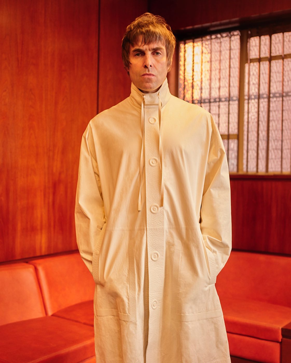 Indoor portrait of a man with a neutral expression wearing a cream-colored coat in front of an orange wall with a modern window.