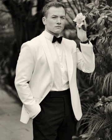 Taron Egerton in a white tuxedo jacket and black bow tie holding a flower outdoors in a portrait by Gavin Bond.