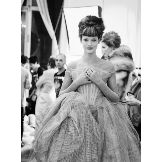 Black and white image of a woman in an off-the-shoulder ball gown in a backstage area surrounded by other people and equipment.