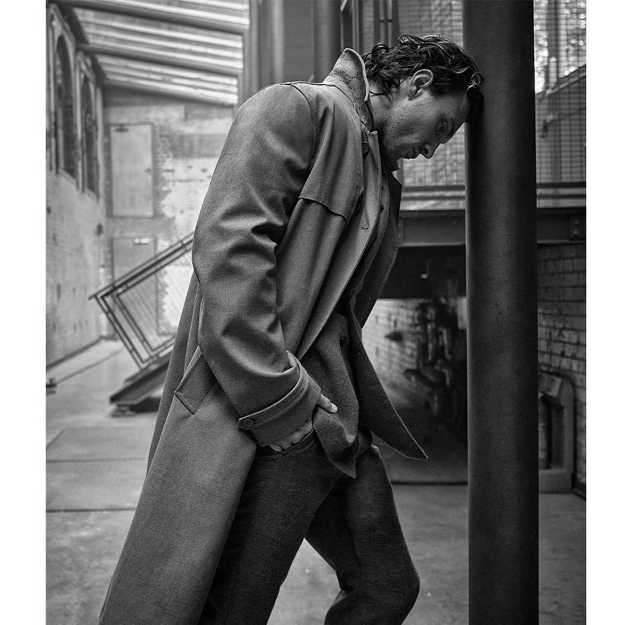 Man in coat stands leaning head against dark metal pole outside in black and white, wearing a coat, pants, and dress shoes.