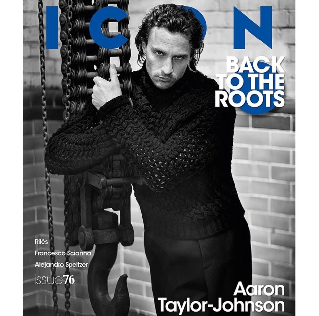 Aaron Taylor-Johnson poses for a magazine cover, wearing a knit sweater and leaning on a rusty, industrial chain-hoist, with a brick wall backdrop.