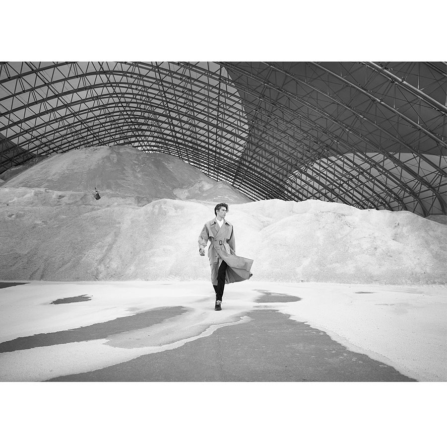 A person in a trench coat walks across a concrete floor toward the camera. Behind them is a large pile of white material under a metal roof.