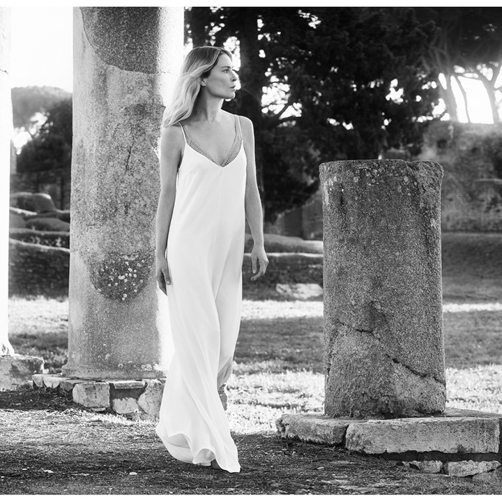 Black and white portrait of an adult woman in a white dress standing near ancient ruins in an outdoor environment with natural lighting.
