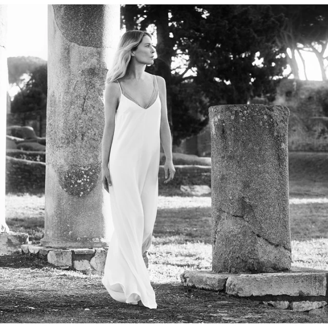 Black and white portrait of an adult woman in a white dress standing near ancient ruins in an outdoor environment with natural lighting.