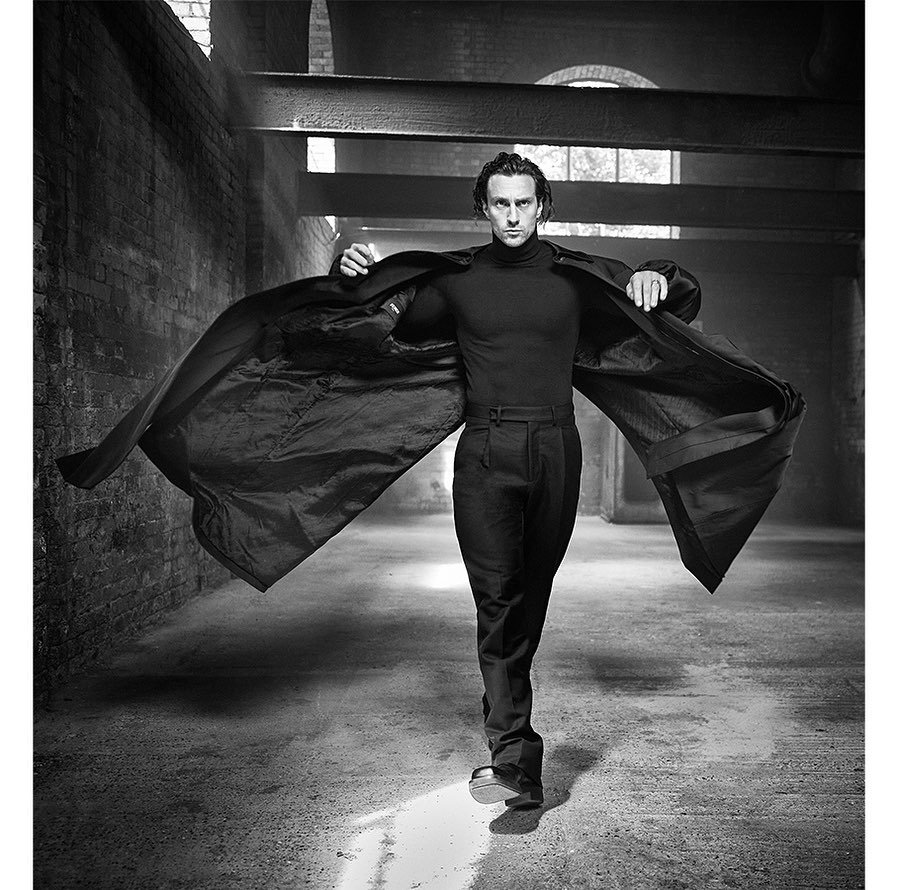 Black and white portrait of a man in dark clothing, wearing an open coat in an industrial hall with natural and artificial lighting.