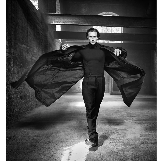Black and white portrait of a man in dark clothing, wearing an open coat in an industrial hall with natural and artificial lighting.