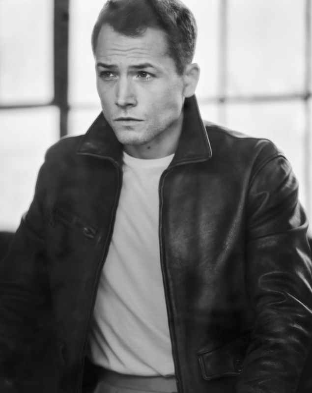Black and white portrait of a man in a leather jacket and white t-shirt looking slightly off-camera with neutral expression and blurred background.