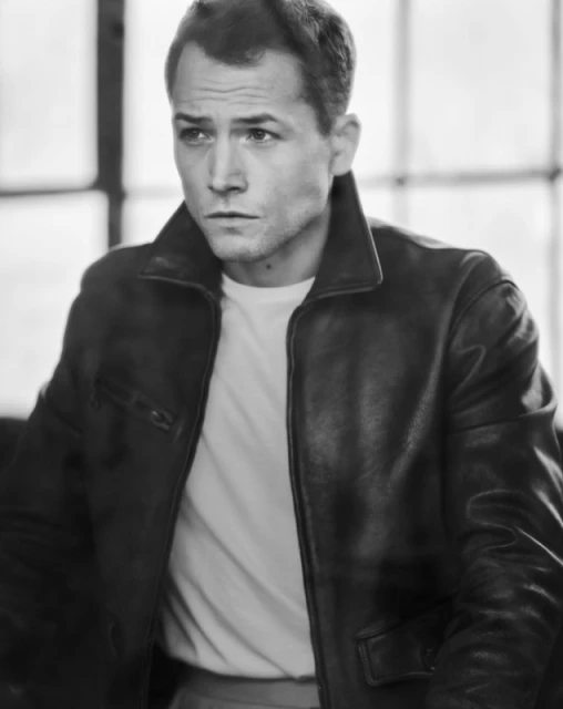 Black and white portrait of a man in a leather jacket and white t-shirt looking slightly off-camera with neutral expression and blurred background.